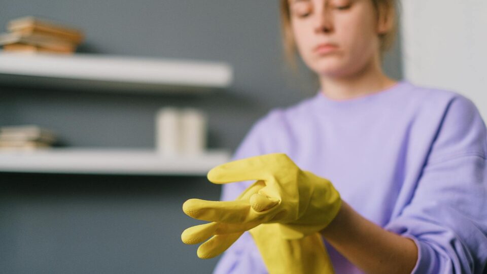 Crop blurred female in casual clothes putting on yellow latex gloves in light room in daytime